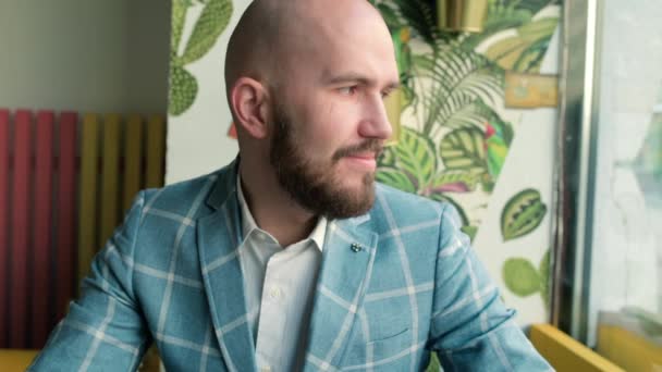 Ein glatzköpfiger Mann mit Bart sitzt in einem Café und schaut aus dem Fenster. Videoporträt — Stockvideo