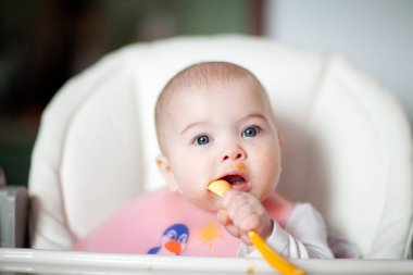 Neşeli bebek kendini kaşıkla yiyor. Yüksek sandalyedeki mutlu kızın portresi..