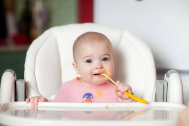 Neşeli bebek kendini kaşıkla yiyor. Yüksek sandalyedeki mutlu kızın portresi..