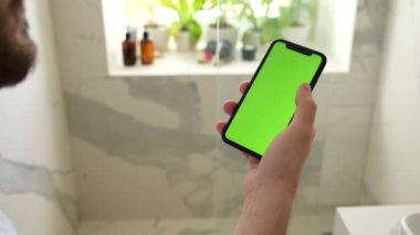 Mans Hand Holding Green Mock-Up Akıllı Telefon 'un yakın çekimi. Modern cep telefonu. Arka plandaki rahat banyoda.