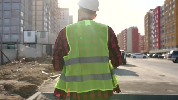 Following Shot of Professional Worker Wearing Hard Hat Holds blueprint, Walking Through Modern constructions site — Stock Video
