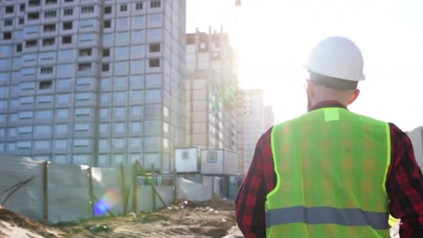 Após tiro de trabalhador profissional vestindo chapéu duro detém planta, andando através de canteiro de obras modernas — Vídeo de Stock