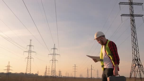 Инженер в белом шлеме проверяет план на фоне линий электропередач — стоковое видео