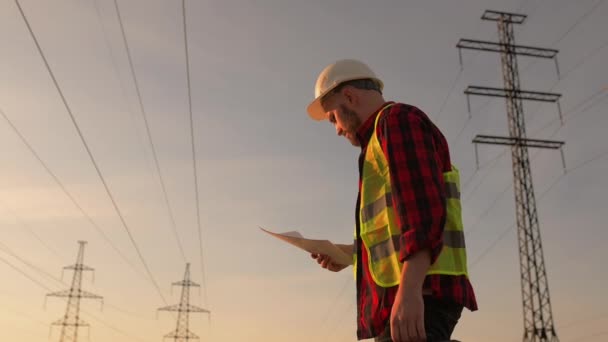 Engineer in a white helmet checks blueprint on the background of power lines — Stock Video