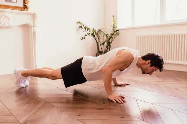 Красивый мускулистый мужчина в футболке и шортах делает функциональные упражнения на полу дома — стоковое фото