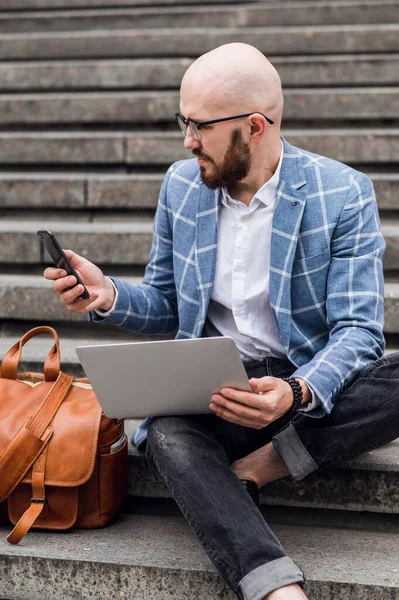 Young bearded businessman sits on steps, using laptop and looks on his screen.Hipster man is working, blogging, chatting online, checking email