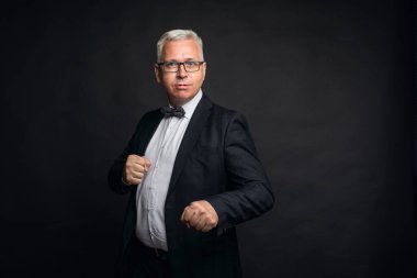 Eksantrik, orta yaşlı, gri saçlı, sıkı takım elbiseli bir adam dövüş pozisyonunda duruyor.