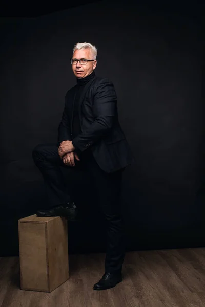 Serious businessman wearing glasses with white hair at the camera with a focused expression against a black studio background