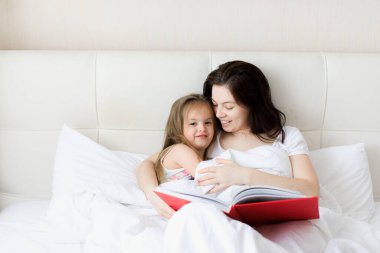 Genç bir anne çocuğuna yatakta kitap okur.