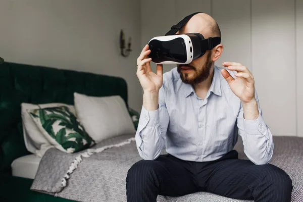 Γενειοφόρος άνθρωπος απολαμβάνοντας γυαλιά εικονικής πραγματικότητας, ενώ κάθεται στο κρεβάτι. Ευτυχισμένος νεαρός με τα ακουστικά σου. — Φωτογραφία Αρχείου