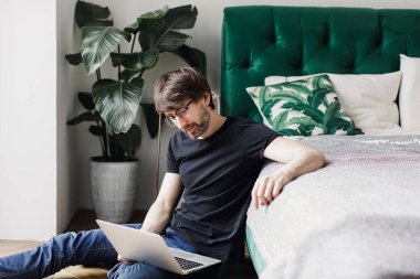  Dizüstü bilgisayarı olan gözlüklü genç bir adam yatağın yanında oturuyor.