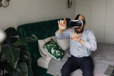 Sanal gerçeklik gözlüklü sakallı genç adam yatağın kenarında oturuyor.