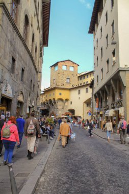 Eski taşlı dar bir sokak dört katlı evler, turist kalabalığı ve mavi gökyüzü.