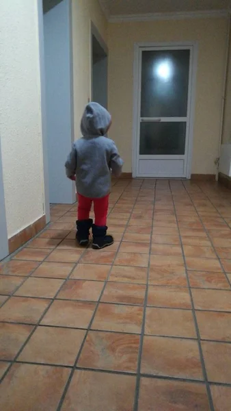A little boy stands in the hallway wearing a gray hooded sweatshirt and red pants