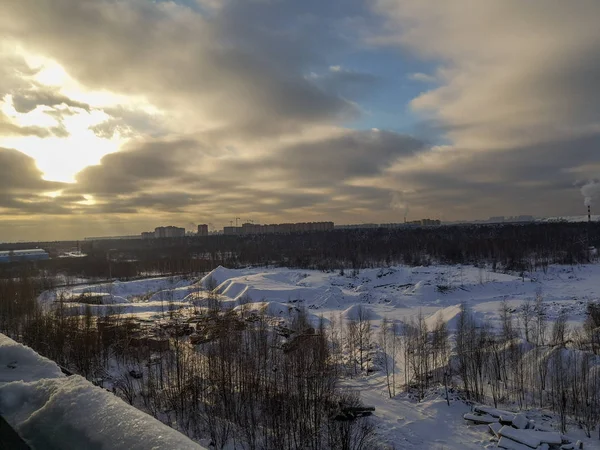 Uzun kar yığınları, çıplak ağaçlar, mavi gökyüzünde alçak gri bulutlar ve ufukta parlayan güneş ve yüksek evler olan bir kış manzarası.