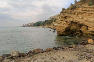 Dağlık kıyı manzarası, kumlu katmanlı taşlar ve kenarda duran evler.