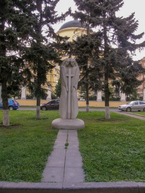 Dini bir heykel. İki kadın başlarını haçın yanına eğmiş. Sarı bir binanın arka planında, siyah çatılı, uzun ağaçlarla çevrili bir bina.