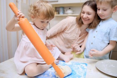 Küçük bir kız pizza hamuru yapmaya yardım ediyor, annesi ve kardeşi ona bakıp gülümsüyor. Seçici odak. 