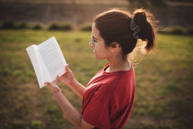 Genç bir kadın, bir parkta gün batımının tadını çıkarırken ve gülümserken güzel bir şeyler okuyor..