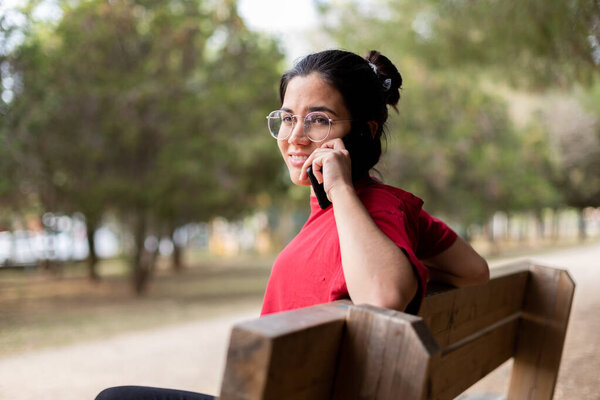 Молодая привлекательная женщина разговаривает по телефону и улыбается в парке, садится на скамейку
