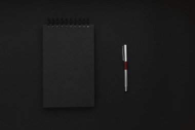 Kapalı, sarmal bir not defteri ve siyah ofis arka planında gümüş kalemi olan minimalist, karanlık bir iş yeri. Minimalizm, iş, serbest meslek kavramı. Boşluğu kopyala.