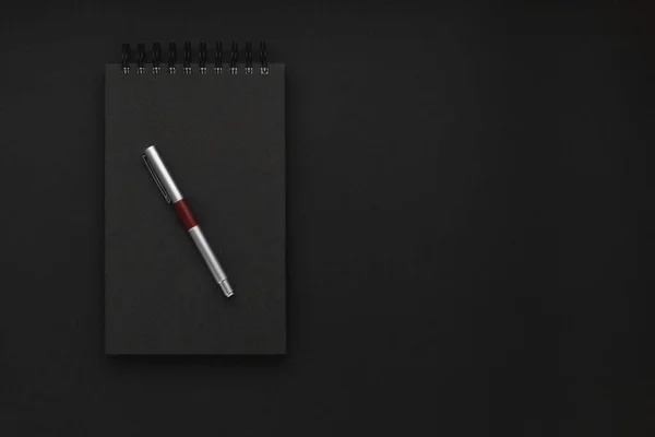 Minimalist dark workplace with a closed black spiral notebook and a silver pen in a black office background. Minimalism, business, freelance concept. Copy space.