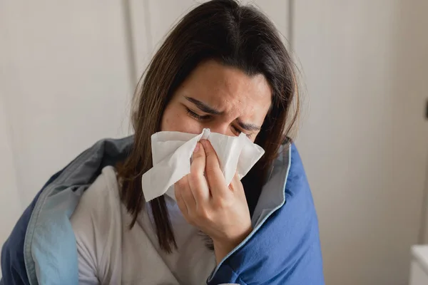 Young ill woman sneezing into a tissue while covering with a blue quilt in her home