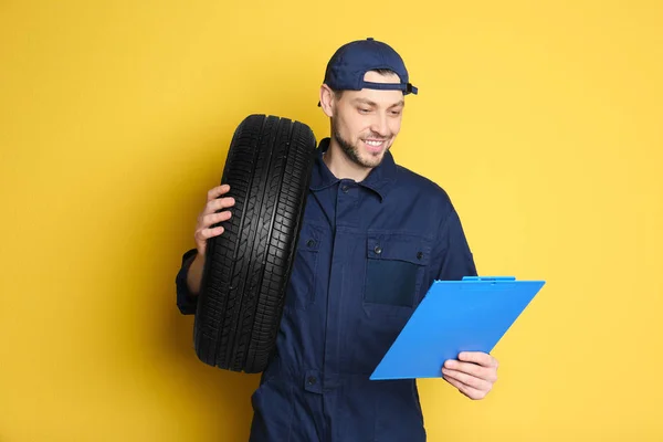 Mecânico masculino em uniforme com pneu de carro e prancheta sobre fundo de cor — Fotografia de Stock