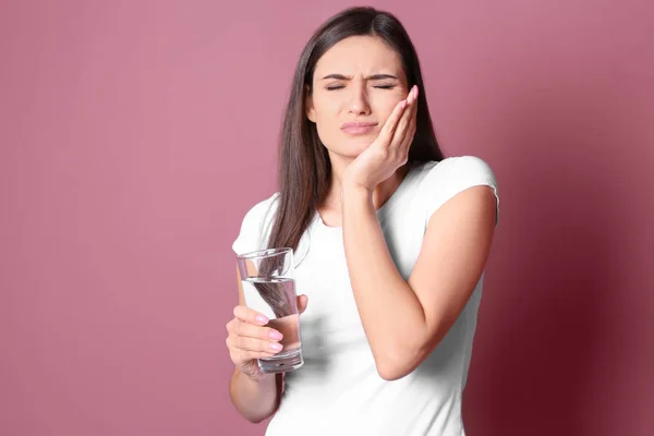 Junge Frau mit empfindlichen Zähnen und einem Glas kalten Wassers auf farbigem Hintergrund — Stockfoto