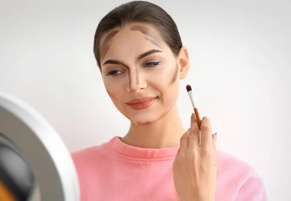 Νεαρή γυναίκα εφαρμόζοντας χάραξη περιγράμματος γραμμές στο πρόσωπό της ενάντια σε ανοιχτόχρωμο φόντο. Προϊόντα επαγγελματικού μακιγιάζ — Φωτογραφία Αρχείου