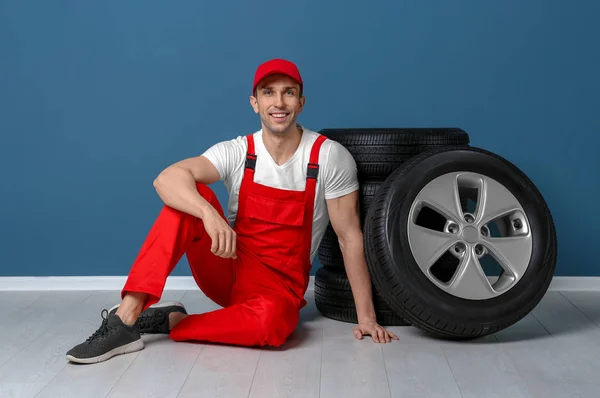 Jovem mecânico sentado perto de pneus de carro perto da parede de cor — Fotografia de Stock