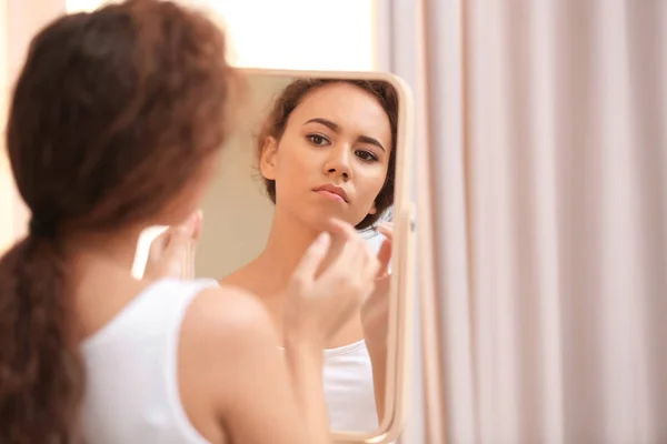 Beautiful young woman with acne problem looking in mirror at home — Stock Photo, Image