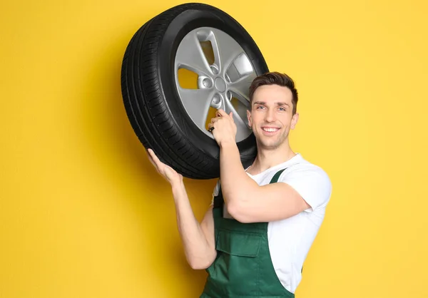 Jovem mecânico em uniforme segurando pneu de carro no fundo de cor — Fotografia de Stock