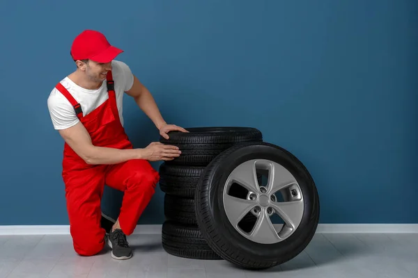 Joven mecánico sentado cerca de neumáticos de coche cerca de la pared de color —  Fotos de Stock