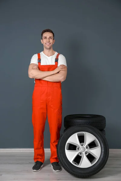 Joven mecánico en uniforme con neumáticos de coche cerca de la pared oscura — Foto de Stock