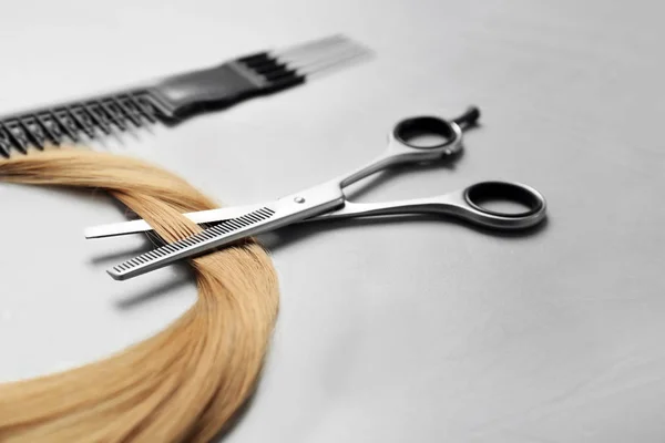 Professional hairdresser scissors, comb and strand of blonde hair on grey background — Stock Photo, Image