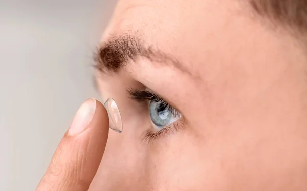 Young man putting contact lens in his eye on light background — Stock Photo, Image