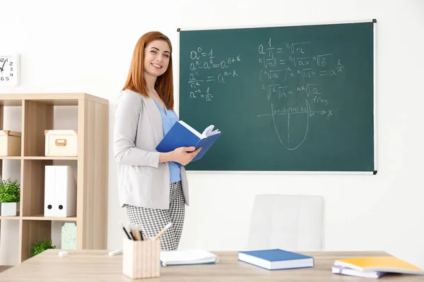 Bela jovem professora explicando fórmulas matemáticas escritas em quadro negro em sala de aula — Fotografia de Stock