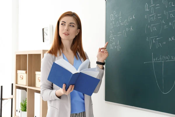 Bela jovem professora explicando fórmulas matemáticas escritas em quadro negro em sala de aula — Fotografia de Stock