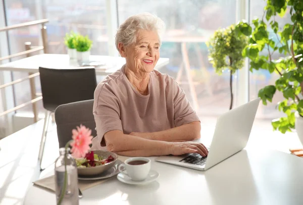 Idős nő használ a laptop otthon a reggeli elfogyasztása közben — Stock Fotó