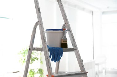 Bucket with paint, brush and gloves on step ladder in light room clipart