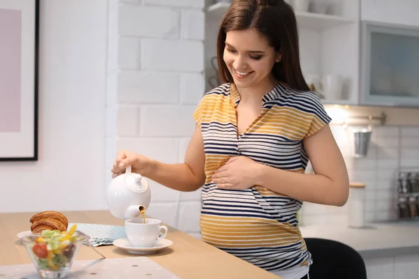 Jovem mulher grávida derramando chá na xícara na mesa na cozinha — Fotografia de Stock