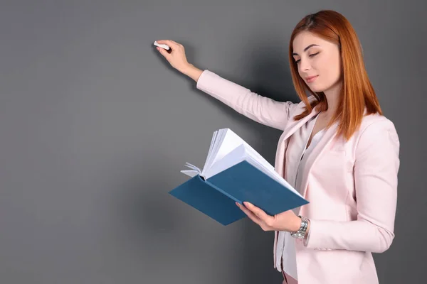 Jovem professora bonita com livro e giz sobre fundo cinza — Fotografia de Stock