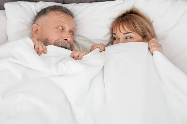 Зрелая пара прячется вместе под одеялом в постели дома — стоковое фото