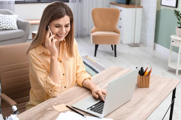Giovane donna che parla al telefono mentre lavora con il computer portatile alla scrivania in home office — Foto Stock