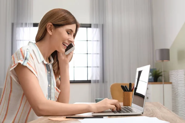 Mujer joven hablando por teléfono mientras trabaja con el ordenador portátil en el escritorio en la oficina en casa — Foto de Stock