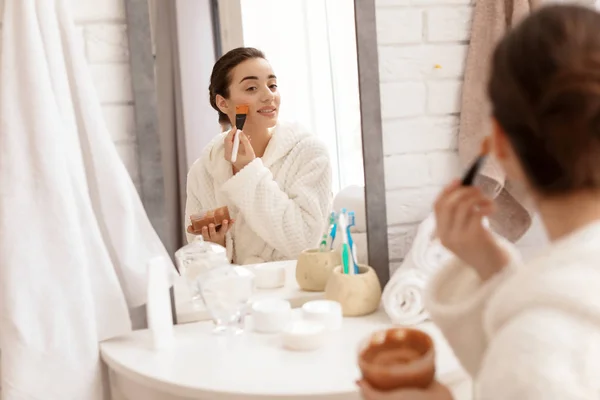 Giovane donna guardando nello specchio e applicando scrub naturale sul viso in bagno — Foto Stock