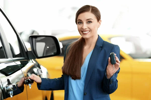 Mujer joven sosteniendo la llave cerca de coche nuevo en el salón — Foto de Stock