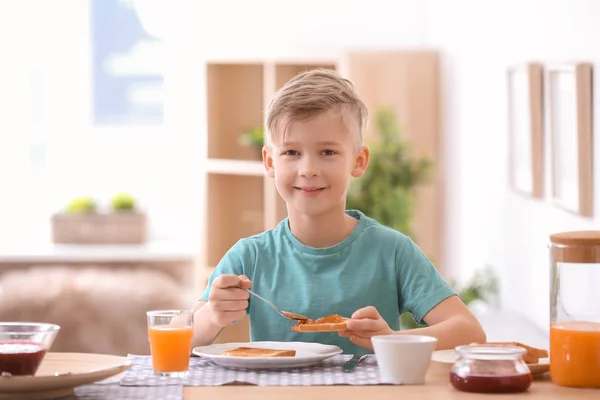 Cute little boy spreading jam onto tasty toasted bread at table