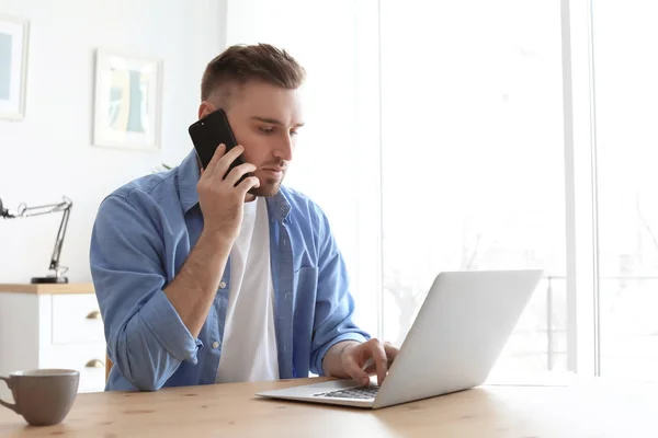 Üstünde hareket eden telefon ile dizüstü bilgisayar masa başında çalışırken konuşan genç adam. Ev ofis — Stok fotoğraf